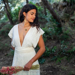 A woman is wearing a lace wedding dress in the forest. The ambiance is dreamy and romantic, and she looks happy. The dress has a boho-chic vibe, perfect for bohemian weddings. The lace of the dress is off-white. The dress has V neck.