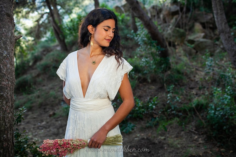 A woman is wearing a lace wedding dress in the forest. The ambiance is dreamy and romantic, and she looks happy. The dress has a boho-chic vibe, perfect for bohemian weddings. The lace of the dress is off-white. The dress has V neck.