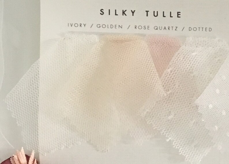 May include: A display card labeled SILKY TULLE featuring four fabric swatches in ivory, golden, rose quartz, and a white dotted pattern. The text on the card lists the colors as IVORY / GOLDEN / ROSE QUARTZ / DOTTED, showcasing the sheer mesh texture.