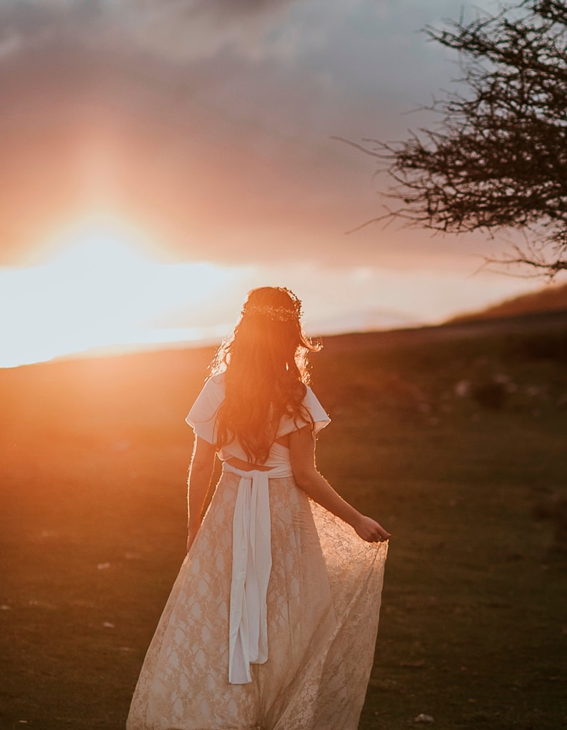 Bride wearing with happiness her boho wedding dress. She is in a landscape that reminds her of Ireland. It's the golden hour and the sun looks incredible.  She is holding the golden lace overskirt of her bridal gown.