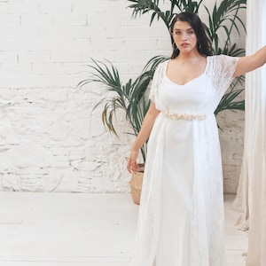 May include: A white lace wedding dress with a fitted bodice and a flowing skirt. The dress has a delicate lace overlay and a simple, elegant design. The model is wearing a gold belt with a floral design.