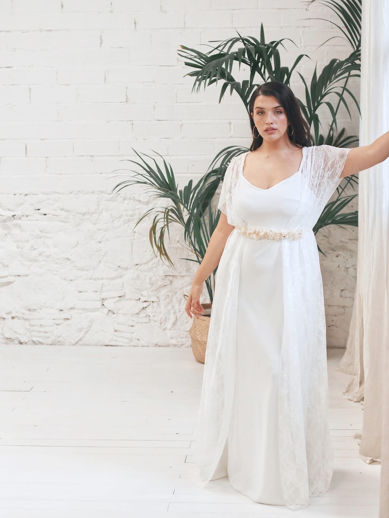 May include: A white lace wedding dress with a fitted bodice and a flowing skirt. The dress has a delicate lace overlay and a simple, elegant design. The model is wearing a gold belt with a floral design.