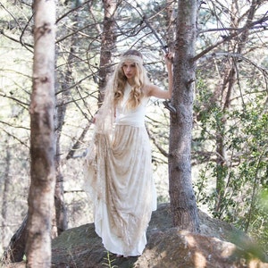Woman in the woods, next to two big trees. She looks boho-chic or hippie while wearing a rustic bridal gown. She has a lace bandana in her hair.