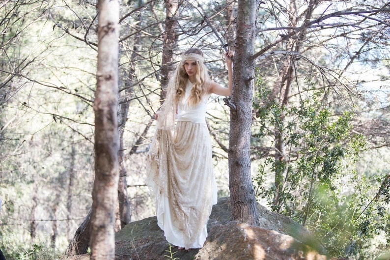 Woman in the woods, next to two big trees. She looks boho-chic or hippie while wearing a rustic bridal gown. She has a lace bandana in her hair.