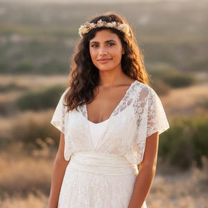 May include: A white lace dress featuring short flutter sleeves, a V-neckline, and a wide waistband. The garment is paired with a floral crown made of dried flowers and foliage. The text www.mimetikbcn.com is visible at the bottom of the image.