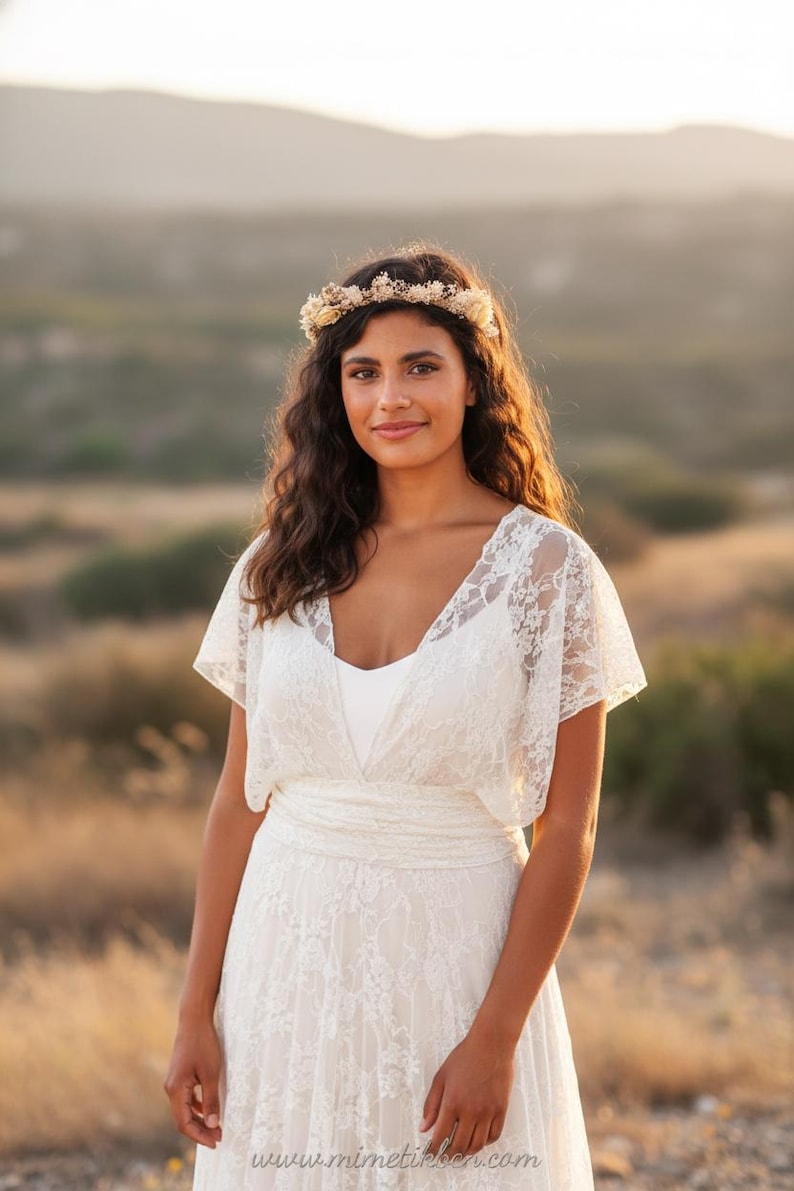 May include: A white lace dress featuring short flutter sleeves, a V-neckline, and a wide waistband. The garment is paired with a floral crown made of dried flowers and foliage. The text www.mimetikbcn.com is visible at the bottom of the image.
