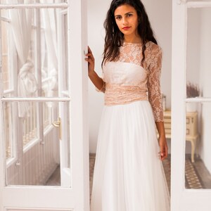 Woman standing in a small and bright room with her hand in a glass door. The room is small but elegant and bright. She is wearing a long sleeve bridal gown that look incredible on her to its convertible design