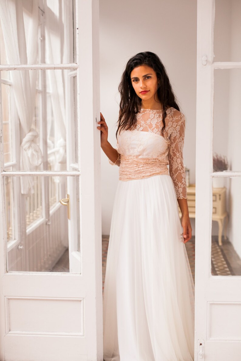 Woman standing in a small and bright room with her hand in a glass door. The room is small but elegant and bright. She is wearing a long sleeve bridal gown that look incredible on her to its convertible design