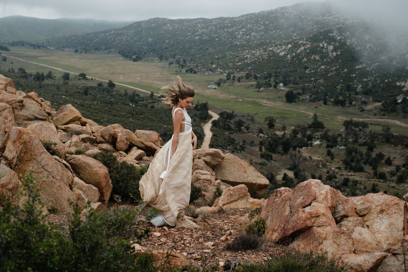 May include: A floor-length dress featuring a white bodice with thin straps and a flowing, textured beige skirt. The garment is worn in an outdoor setting with large rocks and a vast, hilly landscape visible in the background under a cloudy sky.
