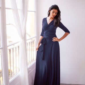 May include: A floor-length navy blue wrap dress featuring three-quarter length sleeves, a V-neckline, and a fabric tie at the waist. The garment is constructed from a solid-colored, smooth material that drapes down to the floor in a full skirt.