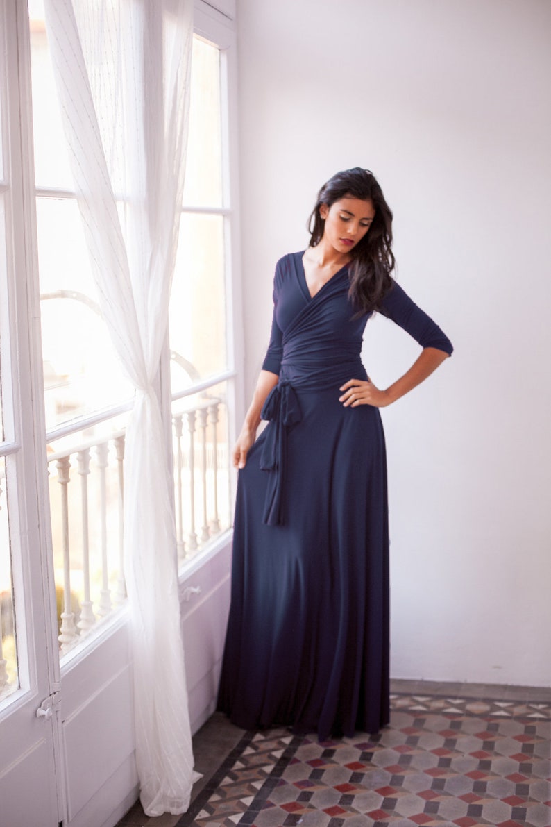 May include: A floor-length navy blue wrap dress featuring three-quarter length sleeves, a V-neckline, and a fabric tie at the waist. The garment is constructed from a solid-colored, smooth material that drapes down to the floor in a full skirt.