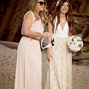 We see a bride and her maid of honor on the beach. They're very happy and smiling. The bride is wearing a long rustic wedding dress. The dress is made in ivory and golden color, creating an elegant contrast. She also wears a big natural bouquet.