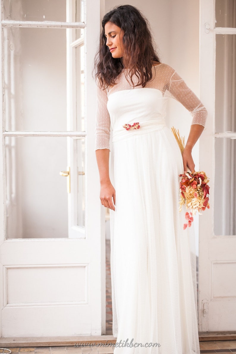 May include: A floor-length white dress featuring a sheer overlay with a subtle dotted pattern, three-quarter length sleeves, and a wide waistband adorned with a cluster of dried red and orange flowers, paired with a matching dried floral bouquet.
