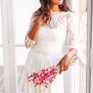 May include: A white lace dress featuring three-quarter length sleeves, a boat neckline, and a gathered waistline. The dress is layered with a floral lace overlay and is held alongside a bouquet of dried pink flowers. The text www.mimetikbcn.com is visible.