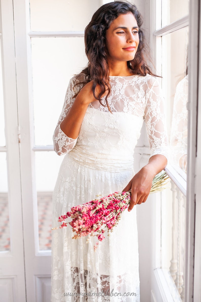 May include: A white lace dress featuring three-quarter length sleeves, a boat neckline, and a gathered waistline. The dress is layered with a floral lace overlay and is held alongside a bouquet of dried pink flowers. The text www.mimetikbcn.com is visible.