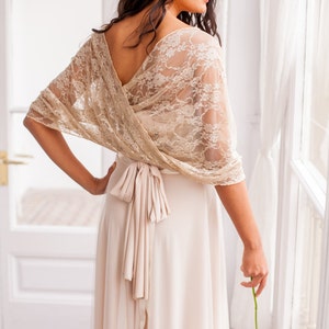 May include: A beige lace bolero wrap featuring a floral pattern and a V-neckline, worn over a solid light beige floor-length dress with a tied waist sash. The lace wrap drapes over the shoulders and arms, creating a layered look with the dress.