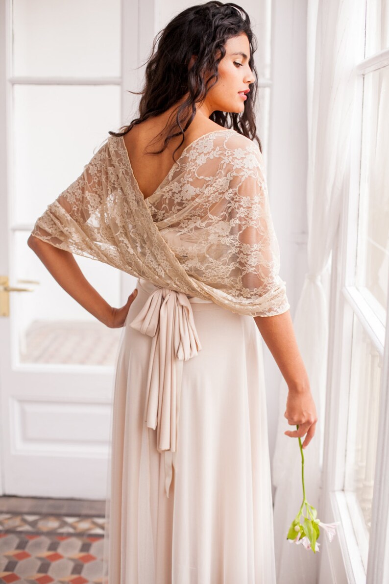 May include: A beige lace bolero wrap featuring a floral pattern and a V-neckline, worn over a solid light beige floor-length dress with a tied waist sash. The lace wrap drapes over the shoulders and arms, creating a layered look with the dress.