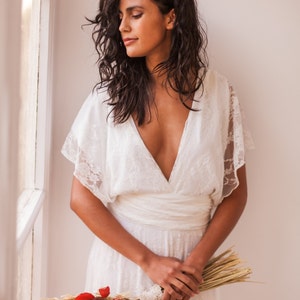 May include: A white lace dress with a deep V-neckline, flutter sleeves, and a wrapped waist detail. The dress features intricate floral lace patterns throughout the fabric. A bouquet of dried red and beige flowers is held in front of the dress.