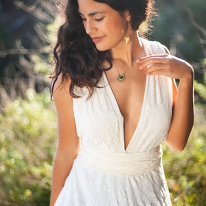 Close up of a woman wearing a wedding dress. She is wearing a boho-chic wedding dress. She wears a V-neck wedding dress. The dress is full made of ivory lace. The woman is also has long dark hair and wears chic bohemian jewelry.