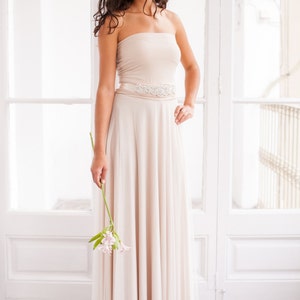 May include: A floor-length strapless dress in a light beige hue, featuring a fitted bodice and a flowing skirt with a sheer overlay. A wide, satin-finish waistband is adorned with a central decorative applique of clear crystals and silver beads.