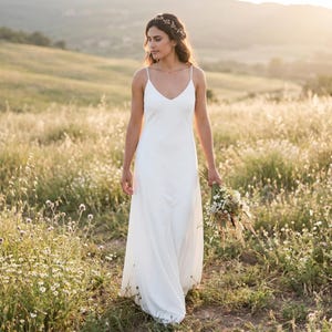 May include: A floor-length white dress with thin spaghetti straps and a V-neckline. The dress features a simple, flowing silhouette and is paired with a delicate floral headpiece and a rustic bouquet of dried wildflowers and greenery.