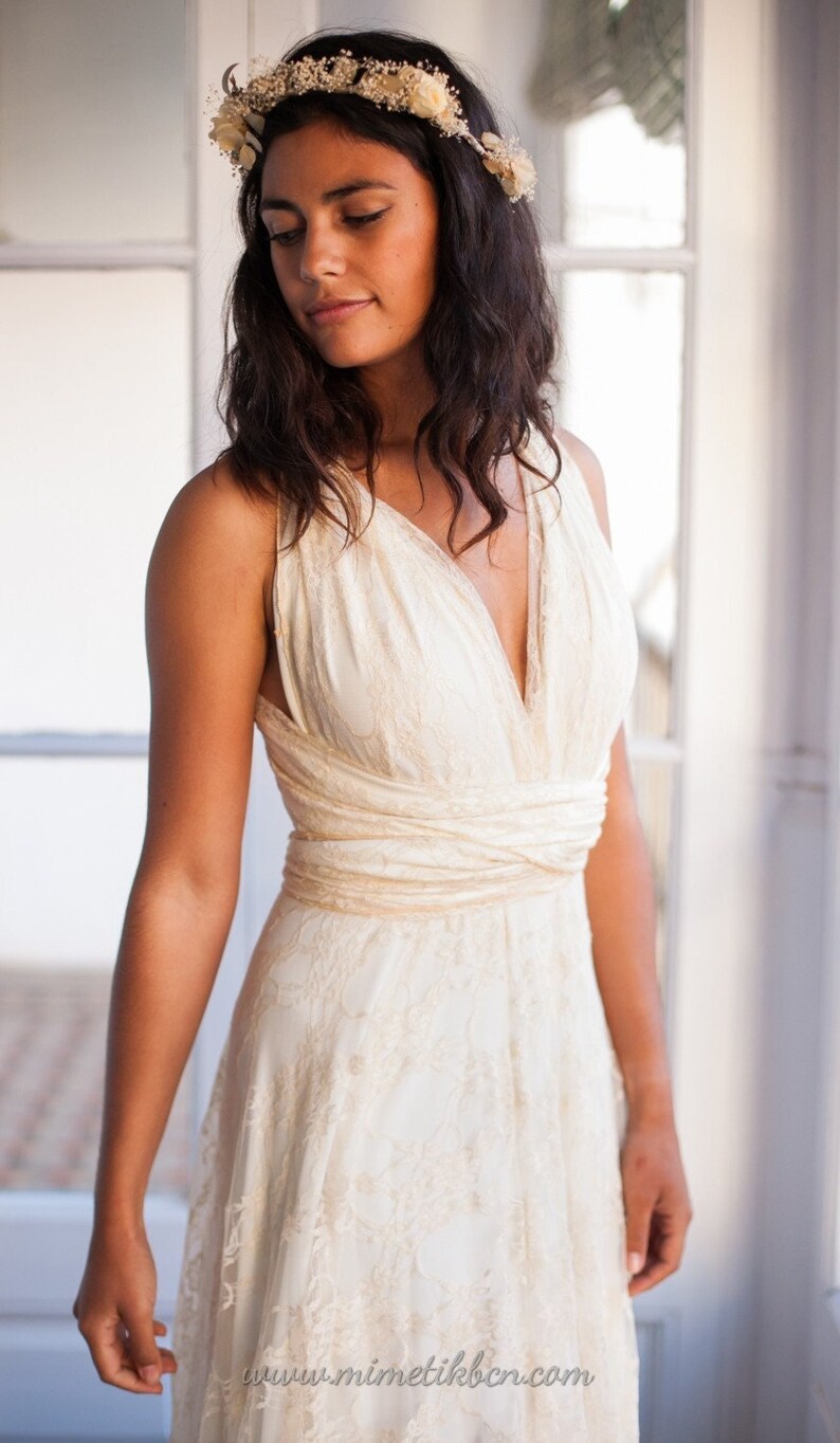 May include: An off-white sleeveless dress featuring a deep V-neckline, a wrapped bodice with horizontal fabric bands at the waist, and an overlay of delicate floral lace. A matching floral crown rests on the head. Text reads www.mimetikbcn.com.