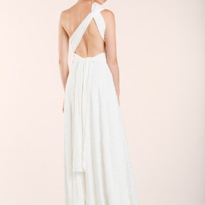 May include: A floor-length white dress featuring a one-shoulder design with wide fabric straps that cross over the open back. The bodice transitions into a long, flowing skirt with a subtle lace texture, creating a continuous silhouette.