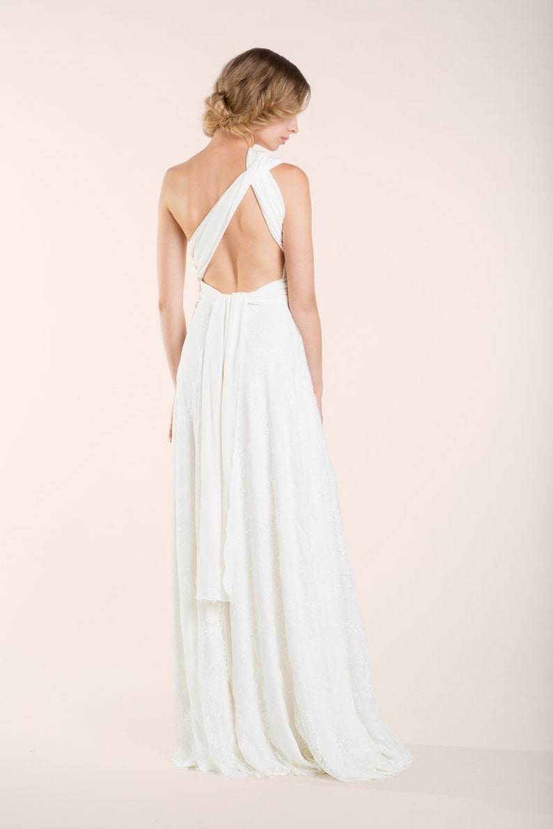 May include: A floor-length white dress featuring a one-shoulder design with wide fabric straps that cross over the open back. The bodice transitions into a long, flowing skirt with a subtle lace texture, creating a continuous silhouette.