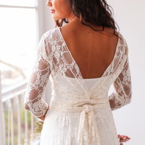 May include: A white lace dress featuring three-quarter length sleeves, a deep V-shaped back neckline, and a matching lace sash tied in a bow at the waist. The garment displays a floral lace pattern over a solid white base layer. Text: www.mimetikbcn.com