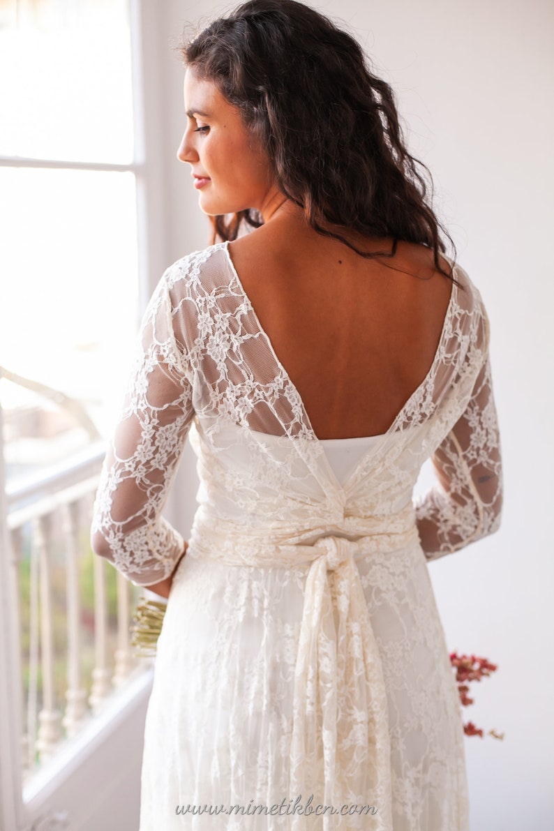 May include: A white lace dress featuring three-quarter length sleeves, a deep V-shaped back neckline, and a matching lace sash tied in a bow at the waist. The garment displays a floral lace pattern over a solid white base layer. Text: www.mimetikbcn.com
