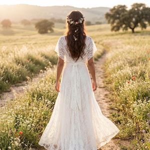 May include: A floor-length white lace dress with short flutter sleeves and a tied waist sash. The dress features a V-neckline and a flowing skirt. The hair is styled in a long braid adorned with a floral crown containing small colorful flowers.