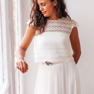 May include: A white lace crop top with a delicate floral pattern and a white tulle skirt. The skirt has a thin belt with a floral embellishment.