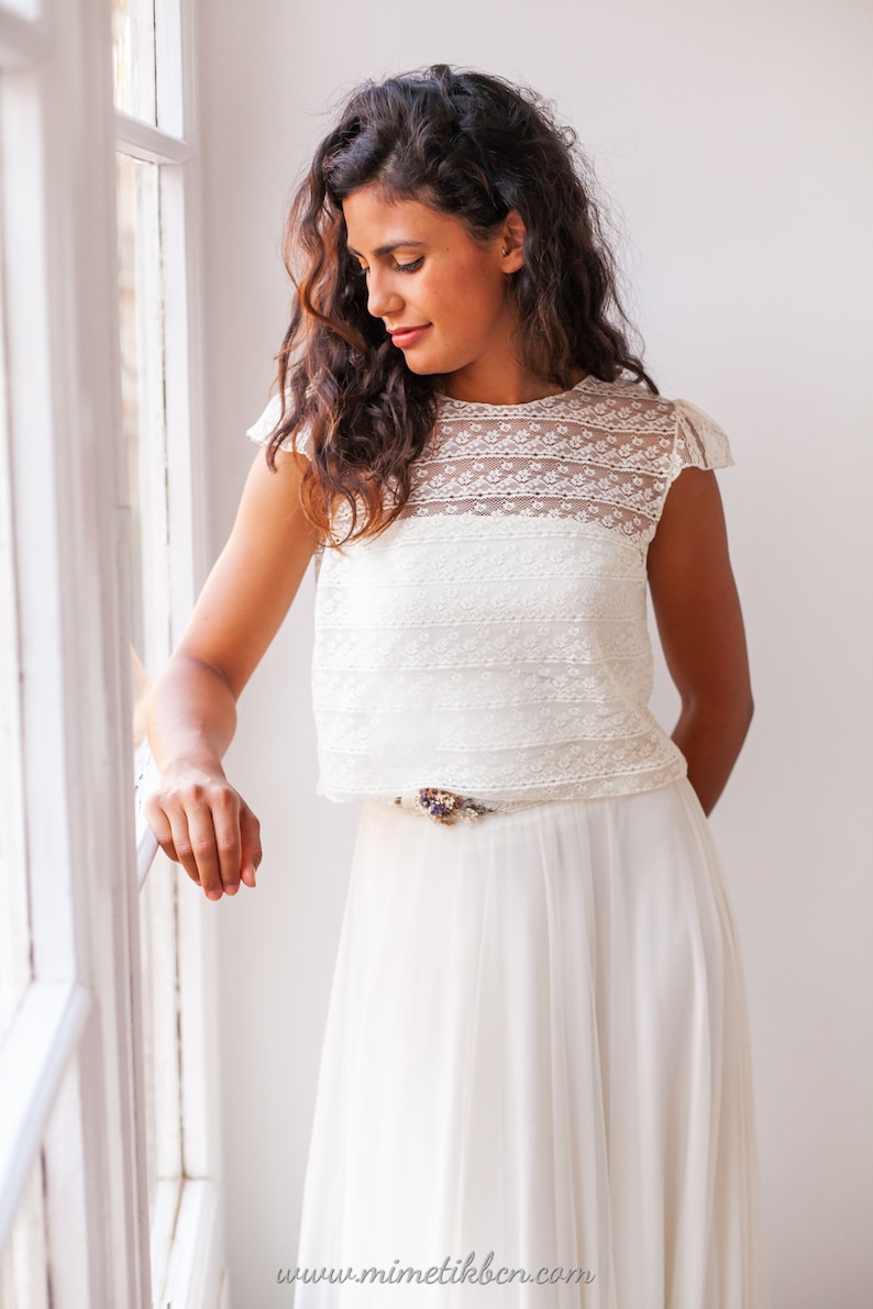 May include: A white lace crop top with a delicate floral pattern and a white tulle skirt. The skirt has a thin belt with a floral embellishment.