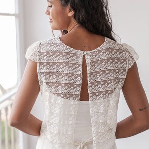 May include: A white lace bridal top with a keyhole back and a tie at the waist. The top is made of delicate lace with a floral pattern.