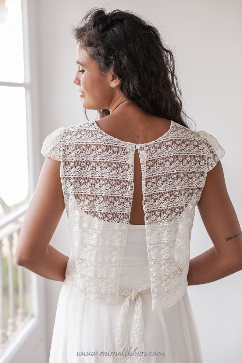 May include: A white lace bridal top with a keyhole back and a tie at the waist. The top is made of delicate lace with a floral pattern.