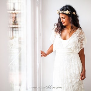 Woman wearing an ivory lace wedding dress near a window. She is looking outside with a dreamy smile. She is brunete and her hair and skin contrasts with her beautiful boho bridal gown