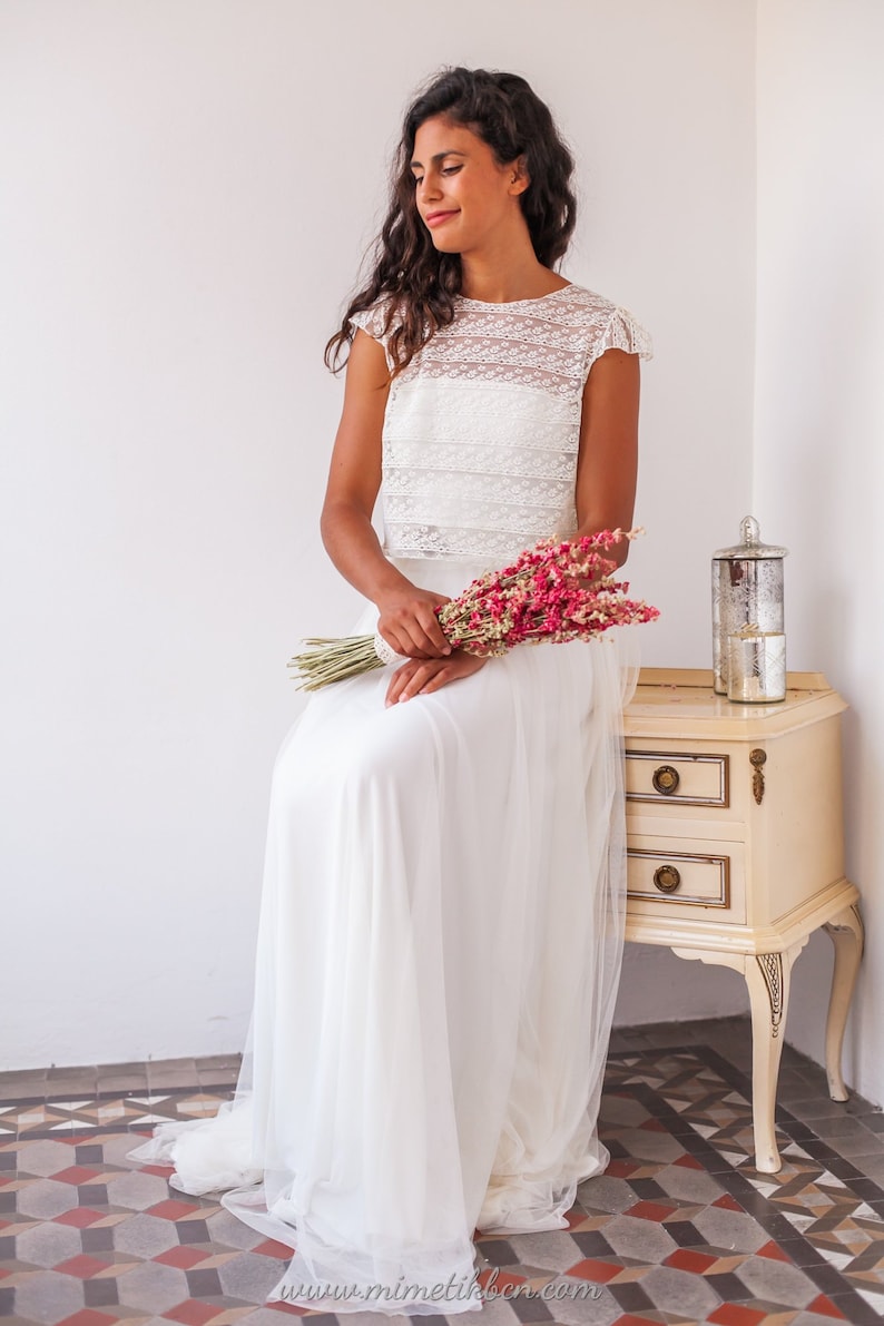 May include: A woman wearing a white lace crop top and a white tulle skirt. She is holding a bouquet of dried pink flowers and is sitting on a white stool in front of a white wall.