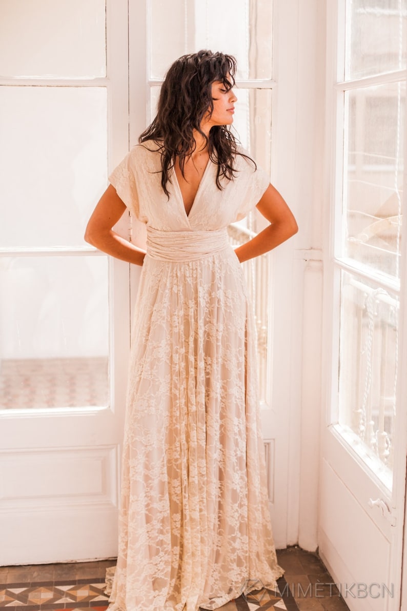 We see a woman next to the window wearing a boho wedding dress. The dress has an ethereal appearence. The woman has dark long hair and is looking to the side. The dress is floor lenght, giving a bohemian and romantic vibe.