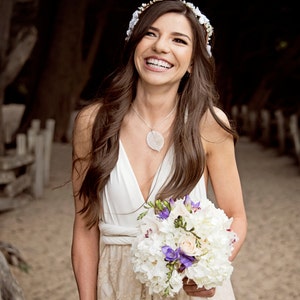 A bride in the beach. She look really happy. She wears a flower crown, a bouquet and has dark hair. She's wearing a rustic wedding dress. The lace skirt of the dress is golden, and the color base is ivory.