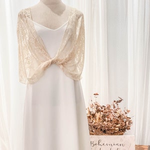 May include: A cream-colored lace bolero with short sleeves and a front knot detail worn over a simple white slip dress. A matching cream-colored headband sits atop the mannequin. A sign reads Bohemian handmade dresses and MIMETIK BCN.