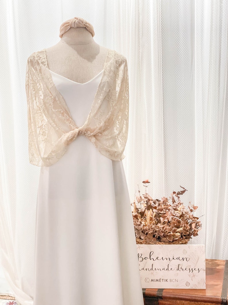 May include: A cream-colored lace bolero with short sleeves and a front knot detail worn over a simple white slip dress. A matching cream-colored headband sits atop the mannequin. A sign reads Bohemian handmade dresses and MIMETIK BCN.