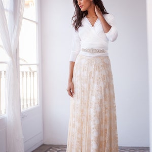 The model is standing up in a clean luminous room. She has a golden lace overskirt and a white bolero.