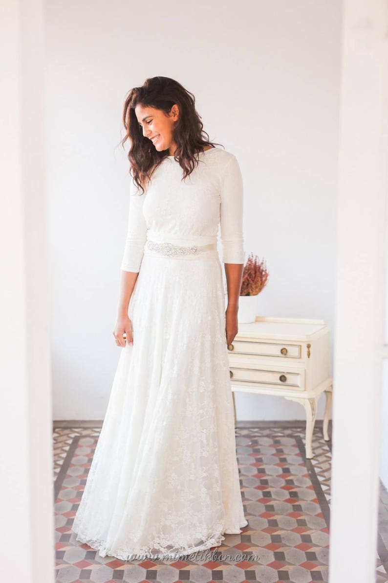 The ivory lace wedding dress looks stunning on the woman. She is a model dressed like a bride. The gown combines a convertible dress with a lace overskirt. She is in front of  drawer