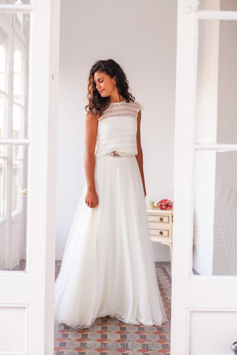 May include: A white lace crop top with a white tulle skirt and a gold belt. The model is standing in a white room with white doors.