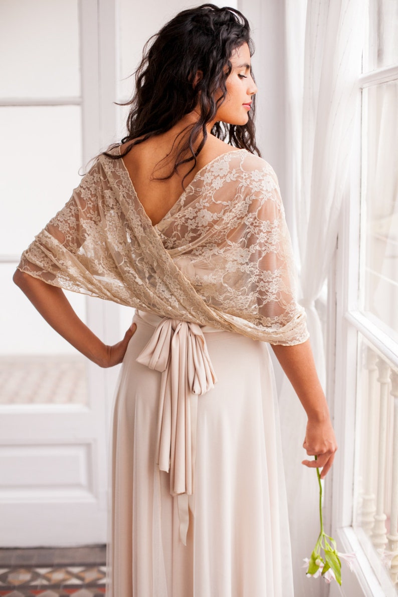 May include: A beige lace shawl draped over the shoulders, featuring a delicate floral pattern and scalloped edges. The shawl is worn over a matching beige dress with a tied waist sash, creating a cohesive look with a soft, neutral color palette.