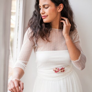 May include: A white dress featuring a sheer overlay with a subtle dotted texture, three-quarter length sleeves, and a round neckline. The waist is accented with a small cluster of dried pink and red flowers. The text www.mimetikbcn.com is visible.