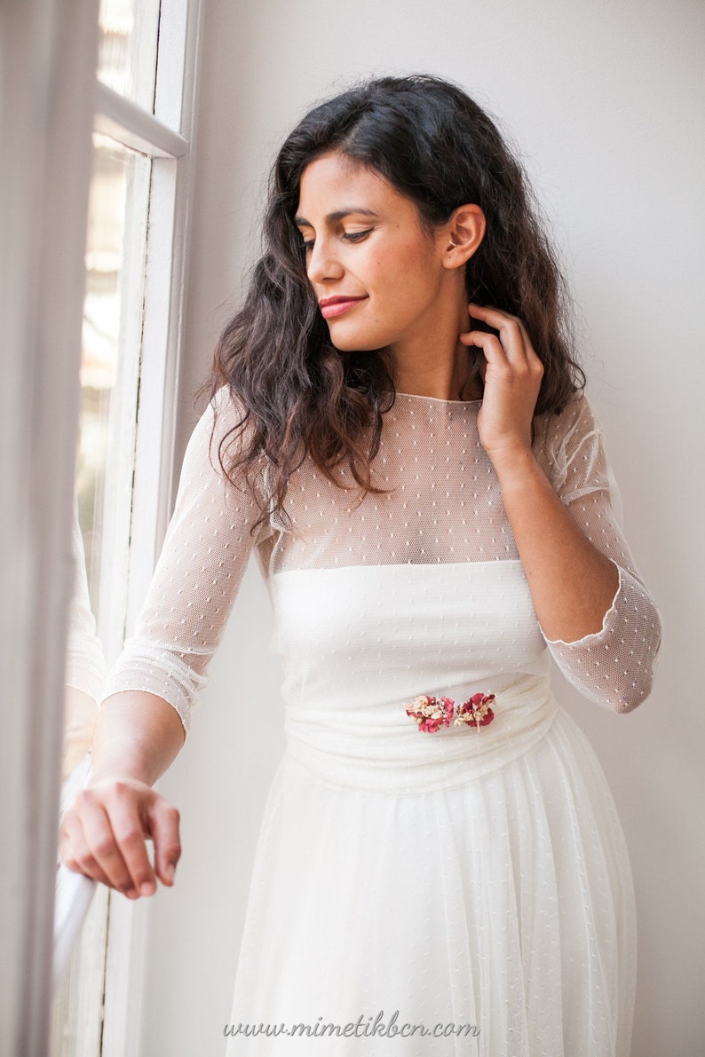 May include: A white dress featuring a sheer overlay with a subtle dotted texture, three-quarter length sleeves, and a round neckline. The waist is accented with a small cluster of dried pink and red flowers. The text www.mimetikbcn.com is visible.