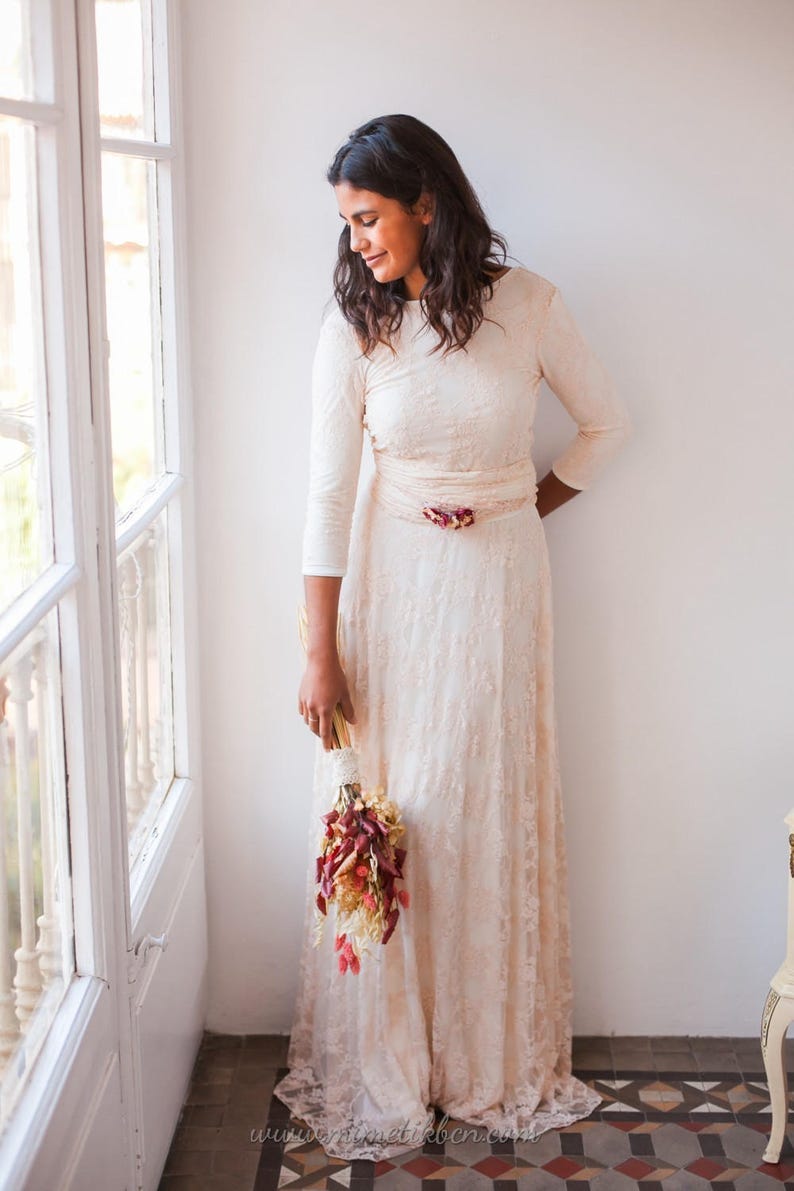 Woman in a bright room staring at the window with a slight smile. She is carrying a bouquet of flowers, that easily combines with the rose quartz lace wedding dress