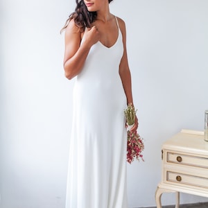 A woman wears a minimal wedding dress. The dress is ivory and has spaghetti straps. She also wears a red flower in her dark hair and a bouquet in her arm.