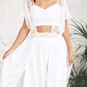 May include: A white lace bridal top with a white crop top underneath and a white flower belt. The top is worn over a white skirt.